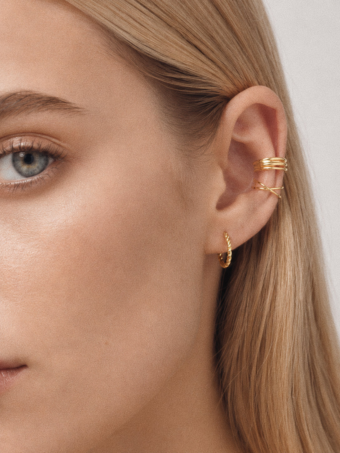 Model wearing a gold criss cross ear cuff and layered cartilage cuffs with a small twisted hoop, styled ear stack look