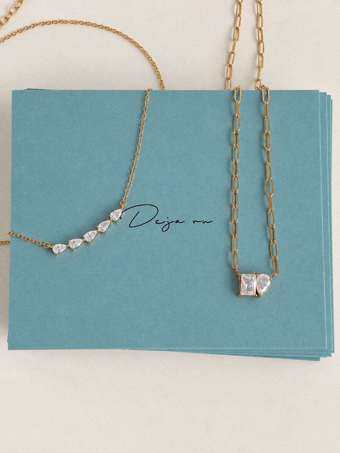 Gold paperclip two stone necklace beside a five-stone necklace on teal Deja vu Jewellery cards.