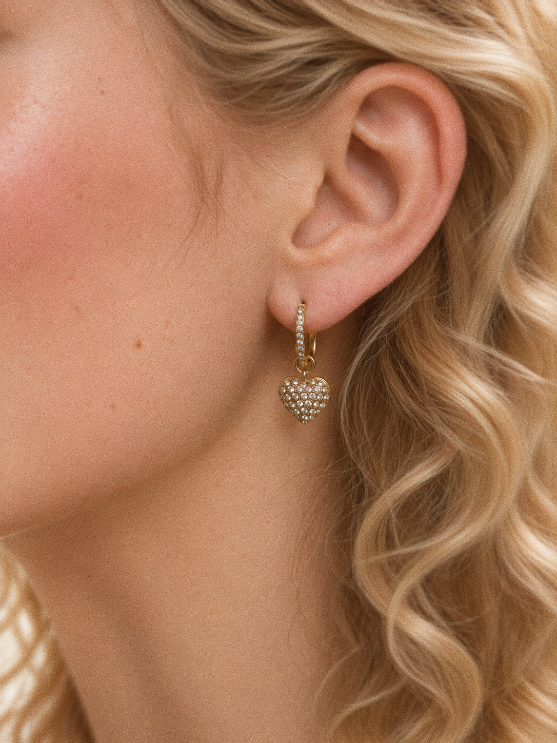 Amour gold heart huggie earring worn on an ear, with a clear-stone heart charm hanging beneath a small decorated hoop.