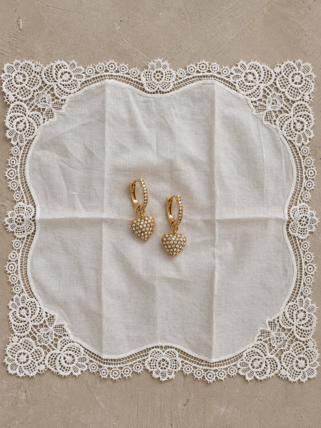 Gold-plated Amour heart charm hoop earrings resting on a white lace-edged cloth, showing the small hoops and sparkling heart drops.