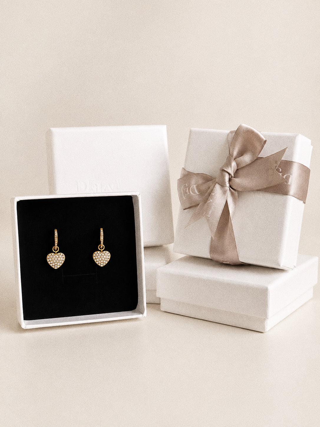 Amour gold heart hoop earrings displayed on black velvet in an open white jewellery box beside boxes with a taupe ribbon.
