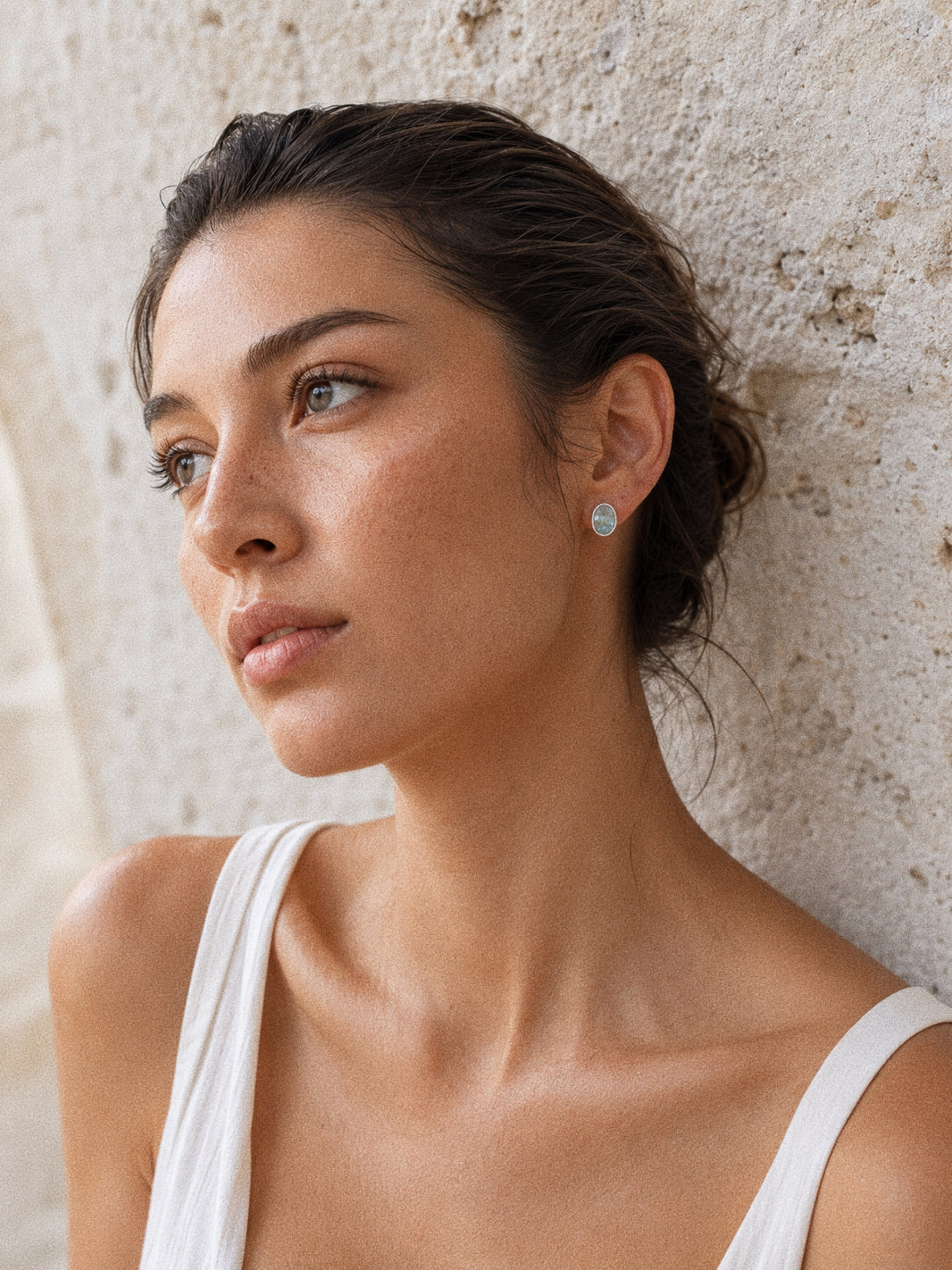 Model in a white top wearing aquamarine cabochon stud earrings against a sunlit stone wall, soft summer light