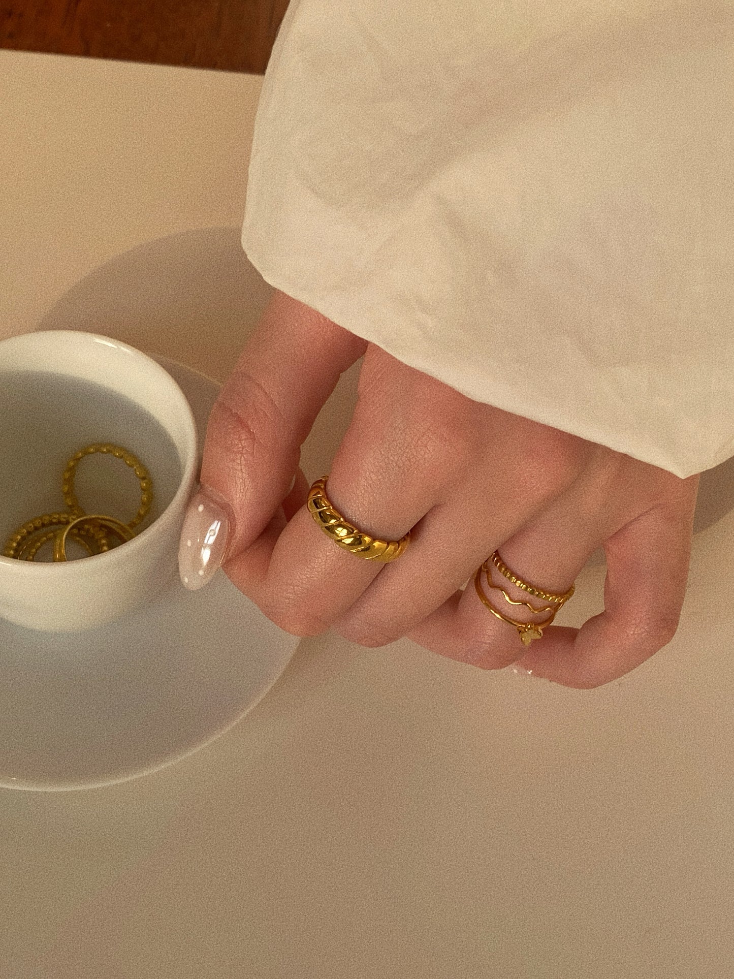 A person wearing a collection of gold titanium steel rings, featuring the wave band, styled next to a white coffee cup and a saucer full of jewellery.