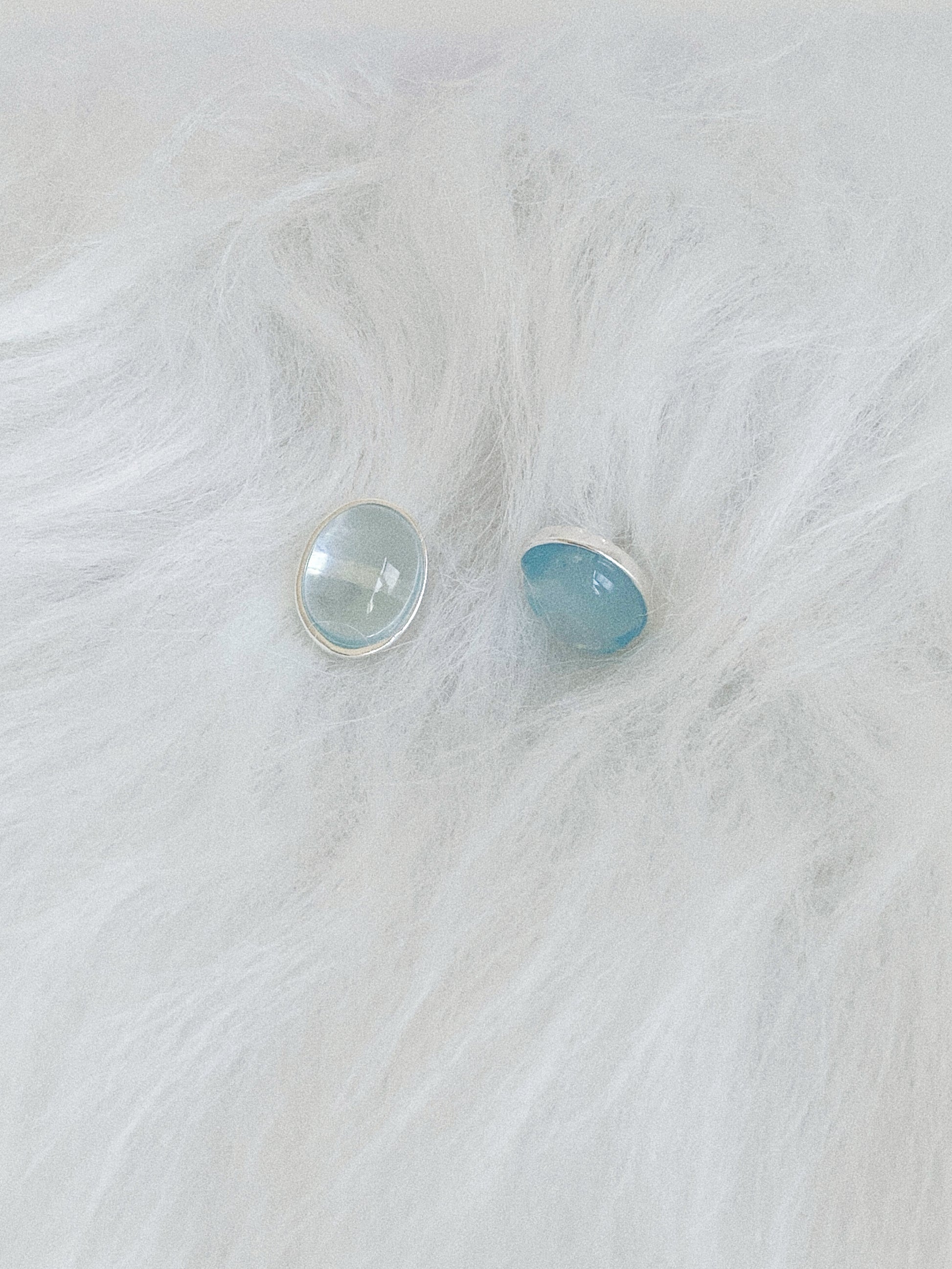 Two aquamarine cabochon stud earrings resting on soft white faux fur, showing their luminous pale blue glow