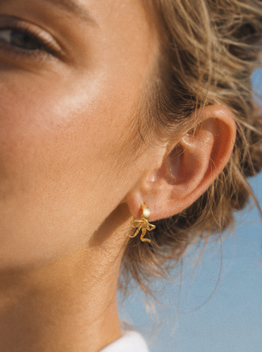 Ballet bow huggie hoop earring in gold worn on the ear, ribbon tails falling just below the lobe.