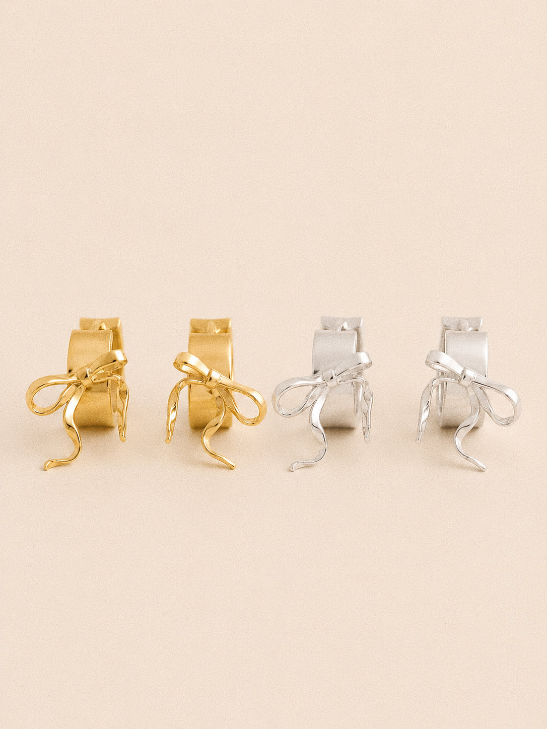 Ballet bow huggie hoop earrings in gold and silver, sculpted ribbon bows with trailing tails on wide hoops.