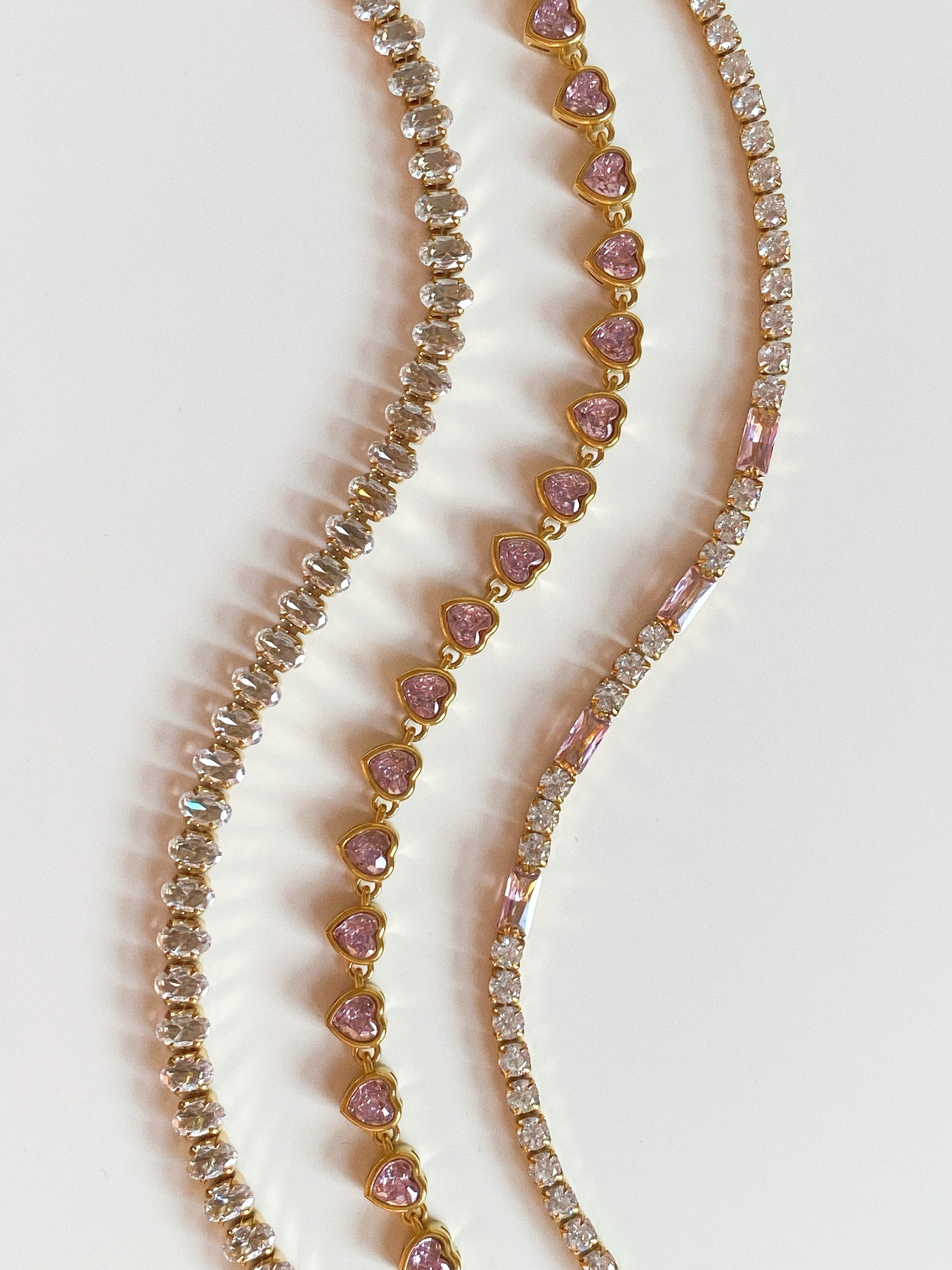 Trio of gold tennis bracelets including pink hearts and mixed cut crystals, flat lay on white background.