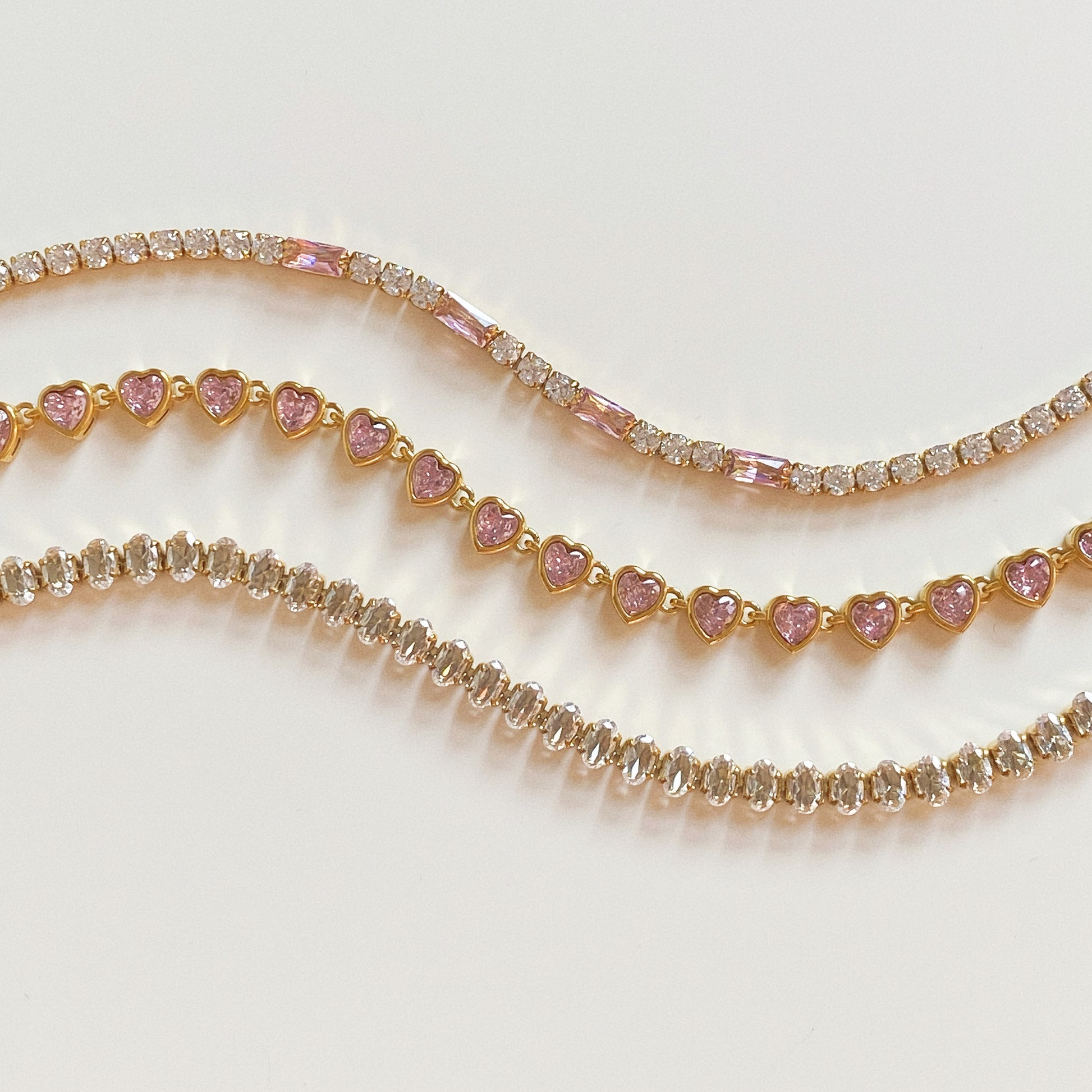 Stack of gold tennis bracelets: Pink Baguette, Pink Heart, and Clear Oval cuts on white background.