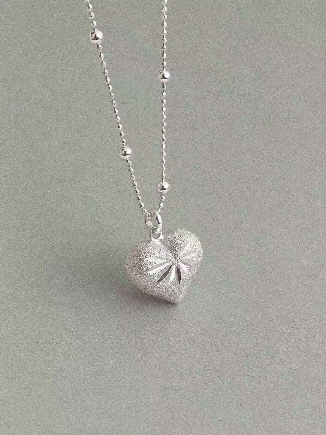 Front view of the rounded silver heart pendant, showing its textured surface and light-catching engraved pattern.