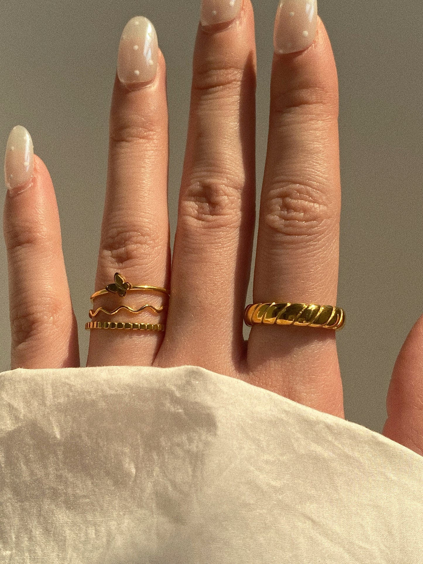A close-up of a hand with neutral-toned nails wearing a stack of three gold rings, including the Mariposa butterfly ring and a wave band.