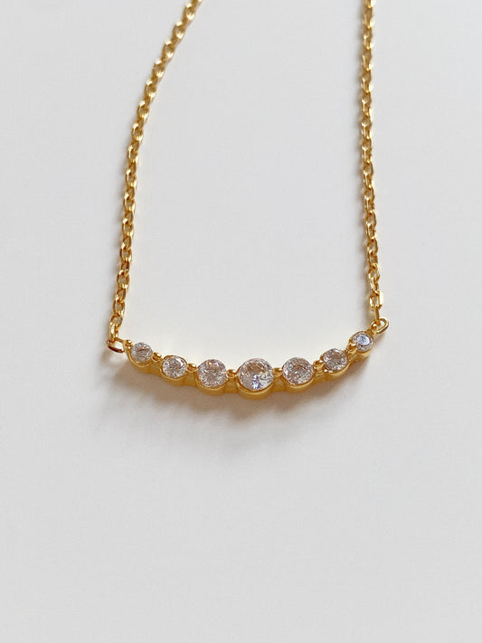 Close up of gold smile necklace featuring a curved bar of seven graduated cubic zirconia crystals.