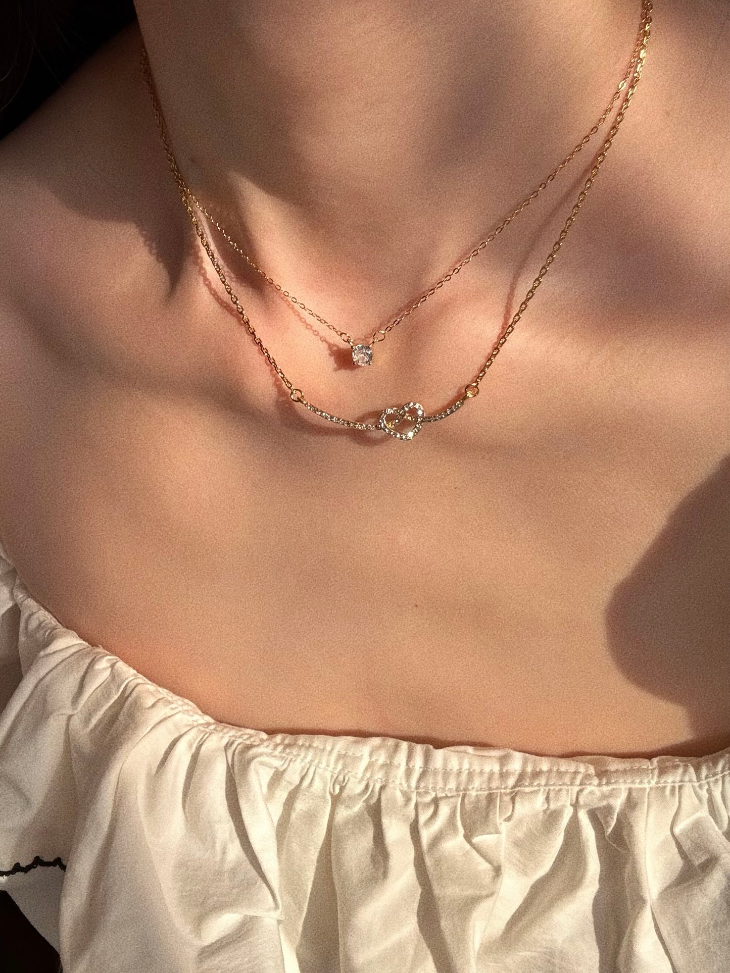 Model wearing the Love Knot Smile Necklace layered with a dainty single crystal solitaire pendant, aesthetic golden hour lighting.