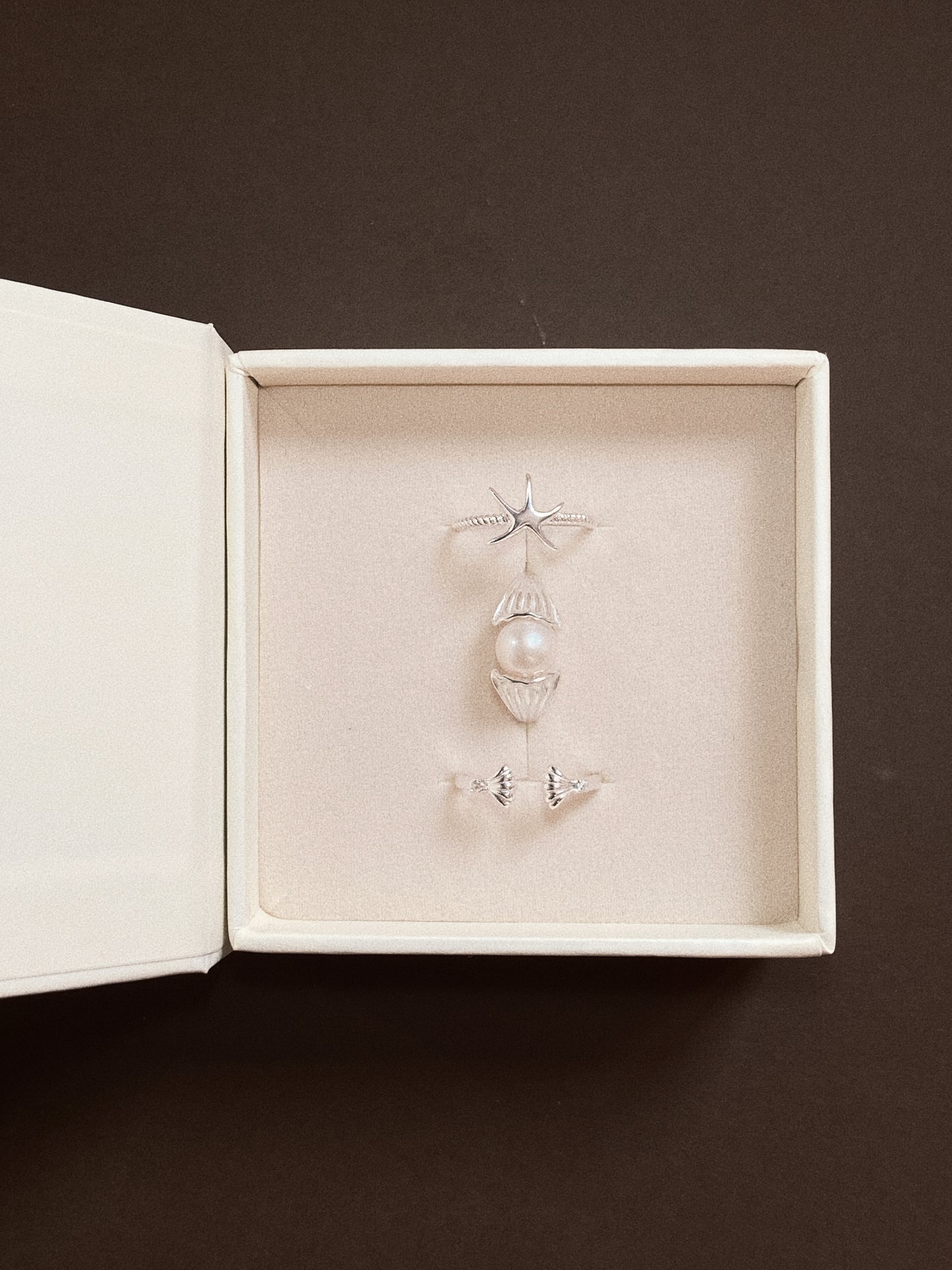 A luxury cream-coloured jewellery gift box containing a set of three silver rings: a starfish ring, a shell ring with a white pearl, and a dainty open shell ring.