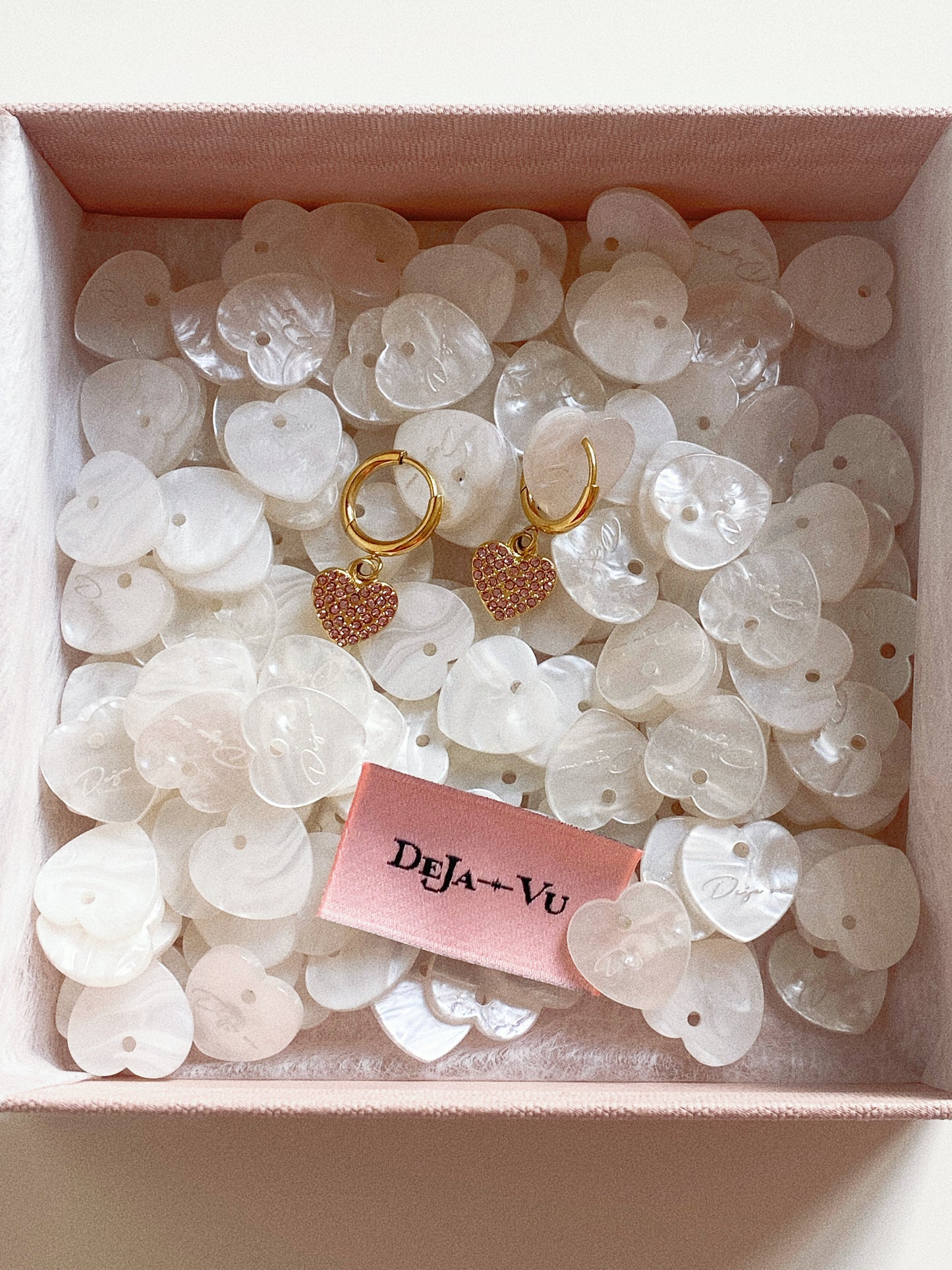 Gold huggie earrings with pink crystal heart charms displayed in a box filled with mother-of-pearl heart shells.