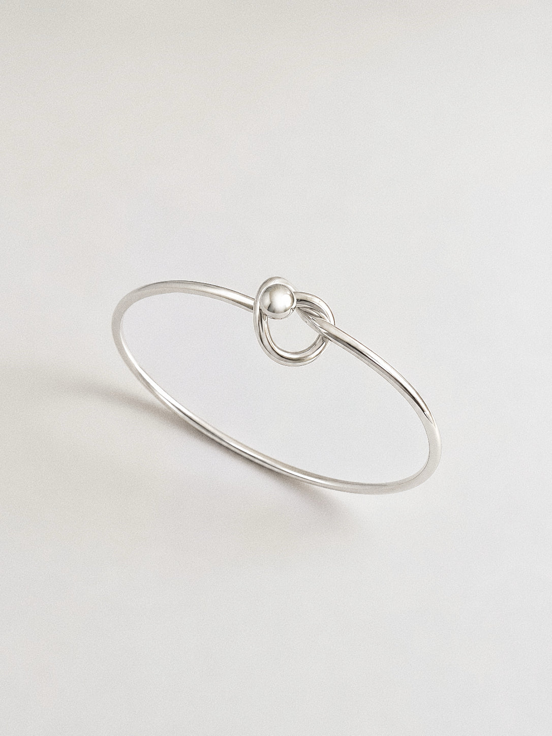 Polished 925 sterling silver love knot bangle with a single sculptural knot and silver ball detail on a soft grey background