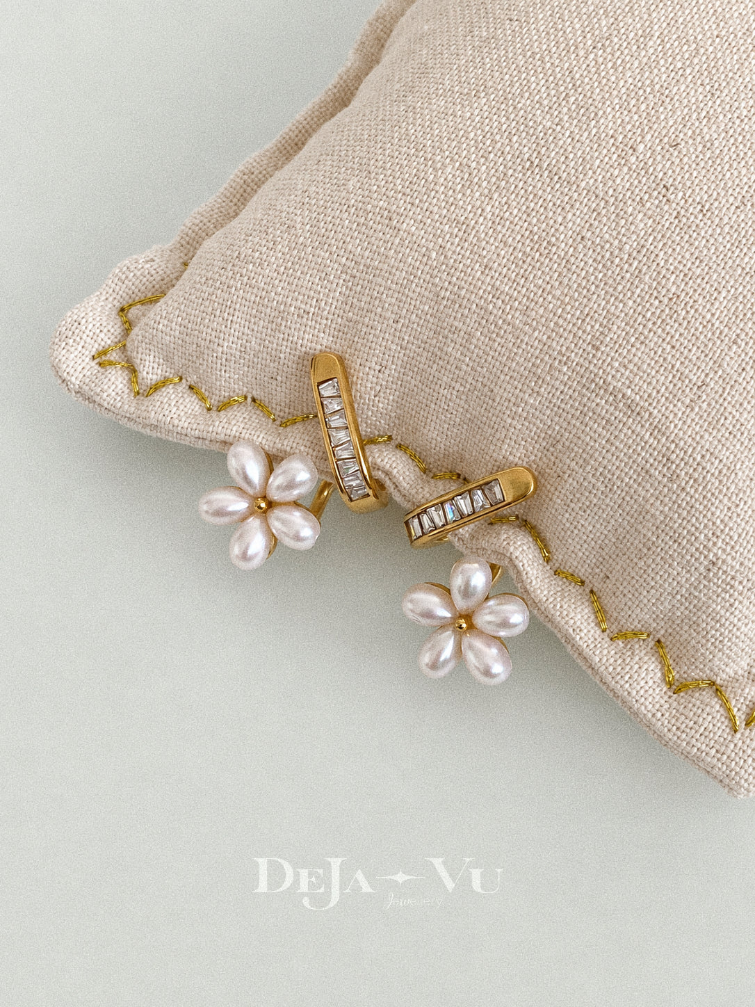 Fleurette faux pearl flower stud earrings resting along a beige linen cushion with gold stitching against a pale green background.