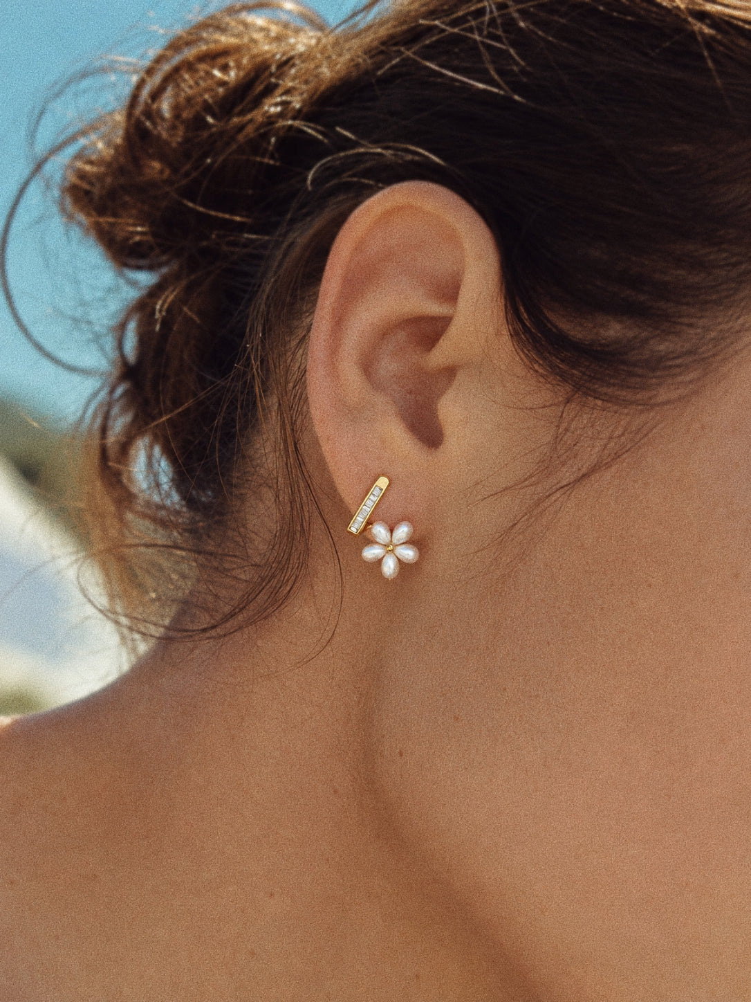 Fleurette flower stud earring worn on an ear, with a slim gold-tone rhinestone bar above a white faux pearl flower.