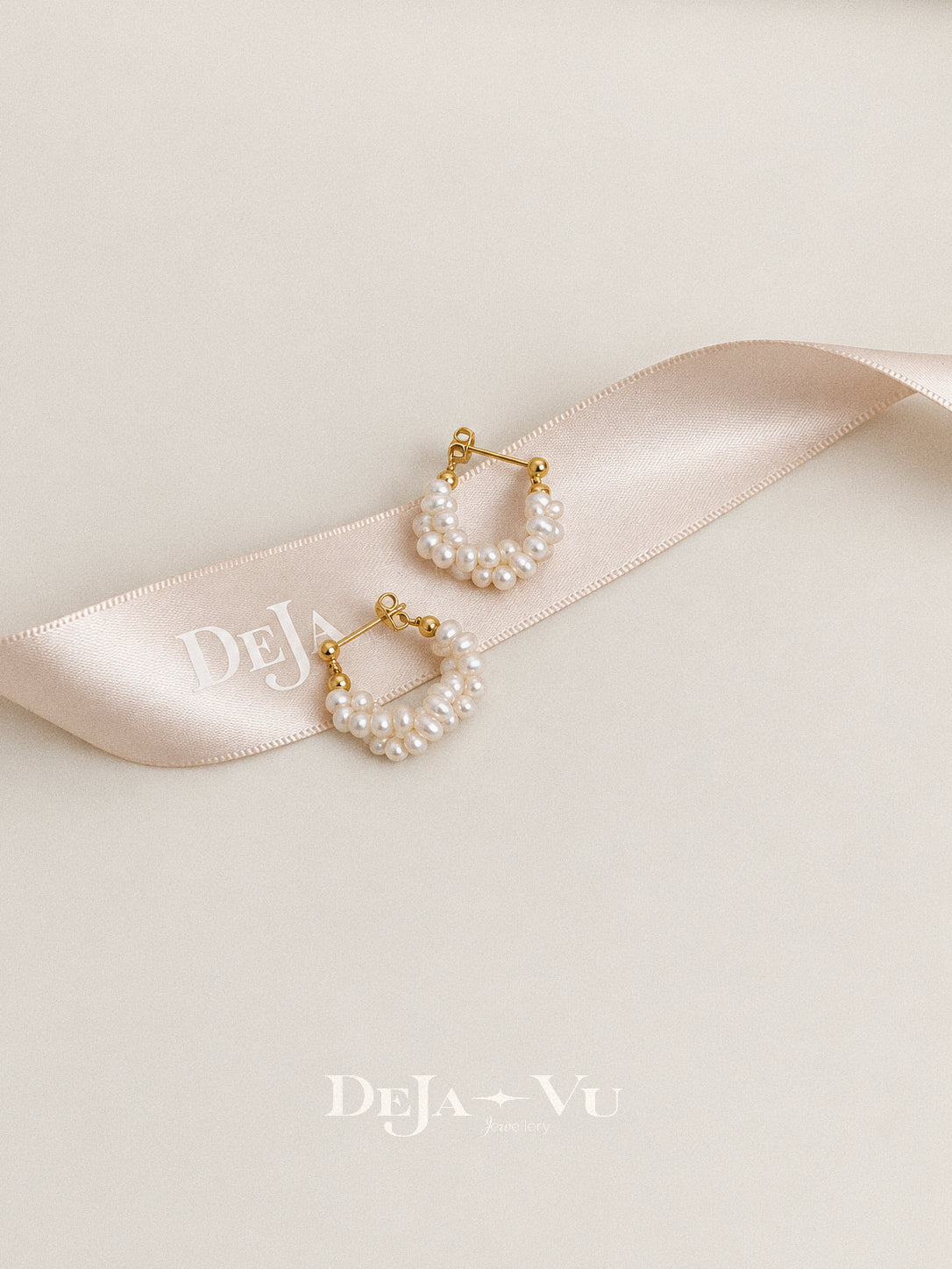 Overhead view of Garland freshwater pearl hoops on a curved branded satin ribbon against an ivory background.