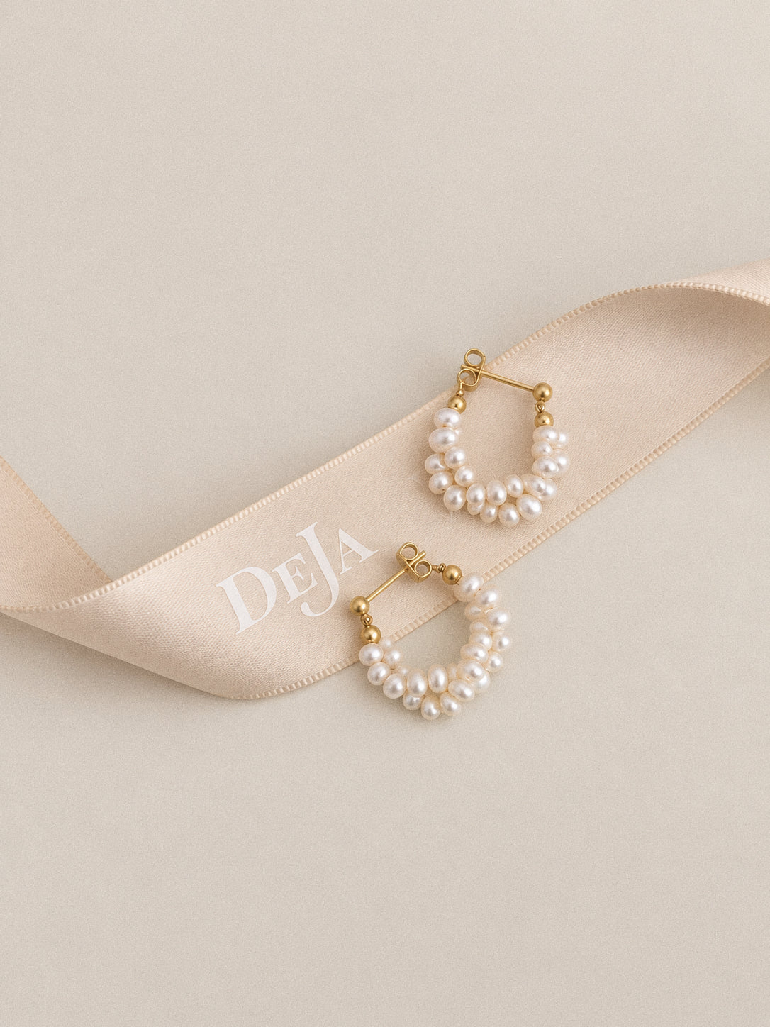Garland pearl hoop earrings arranged on a champagne satin ribbon, showing their twisted wreath-like shape.