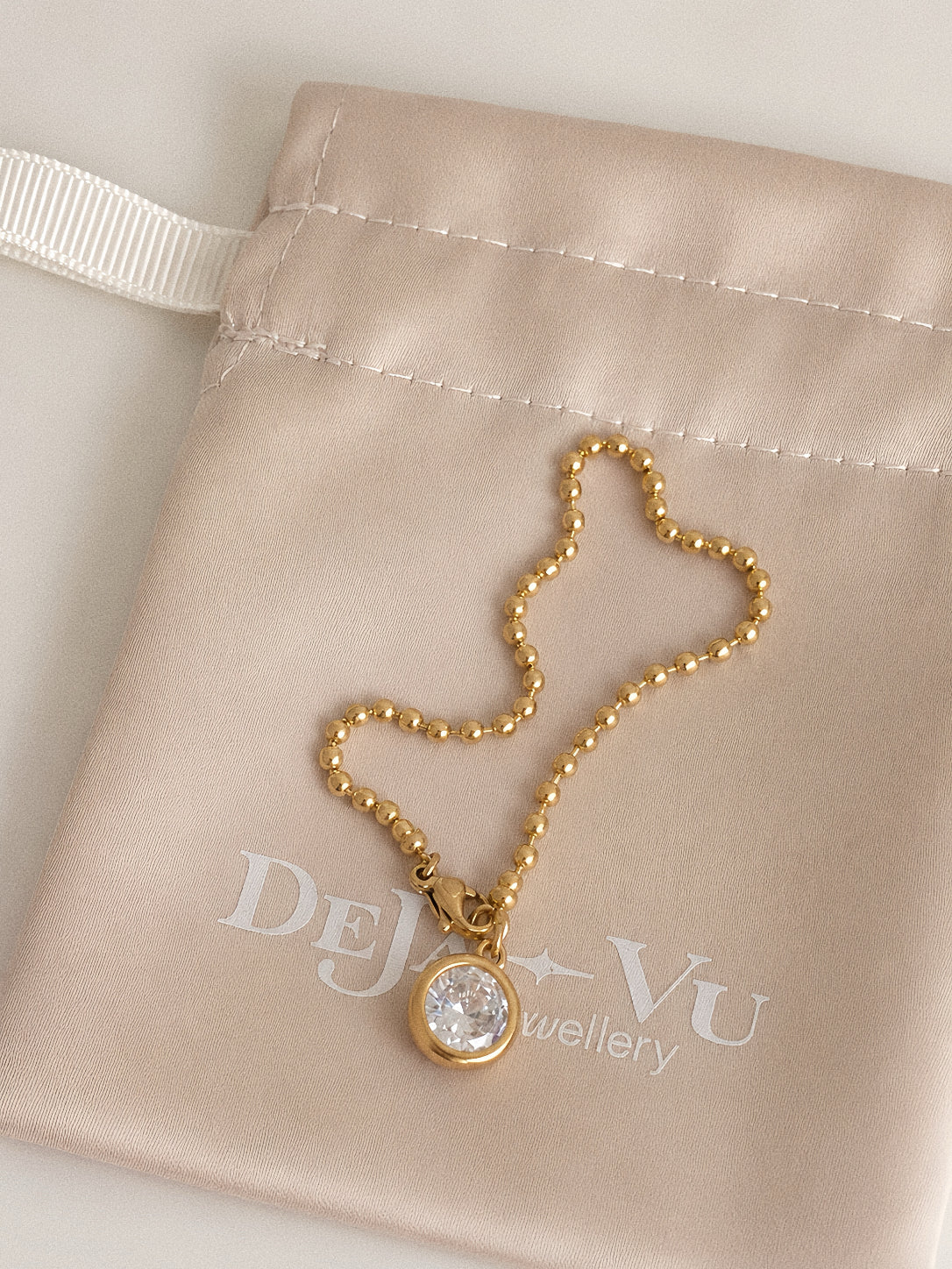 Gold ball chain bracelet with round cubic zirconia charm resting on a Deja Vu Jewellery satin pouch