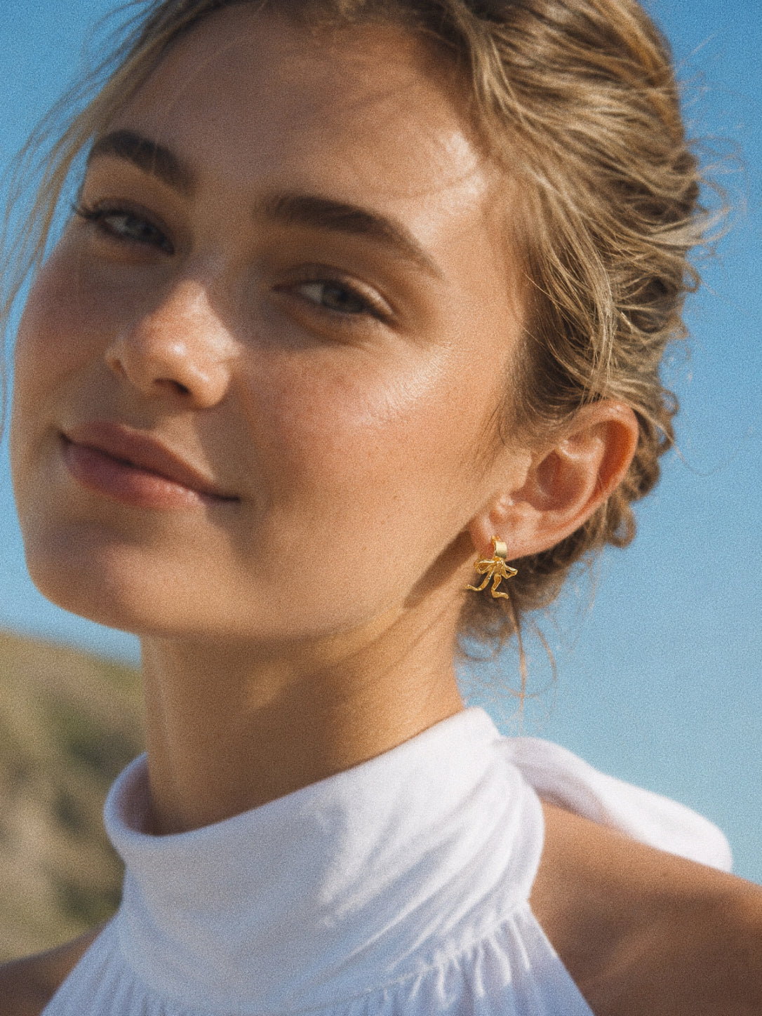 Gold bow huggie earring worn with a white halterneck top against a blue summer sky.