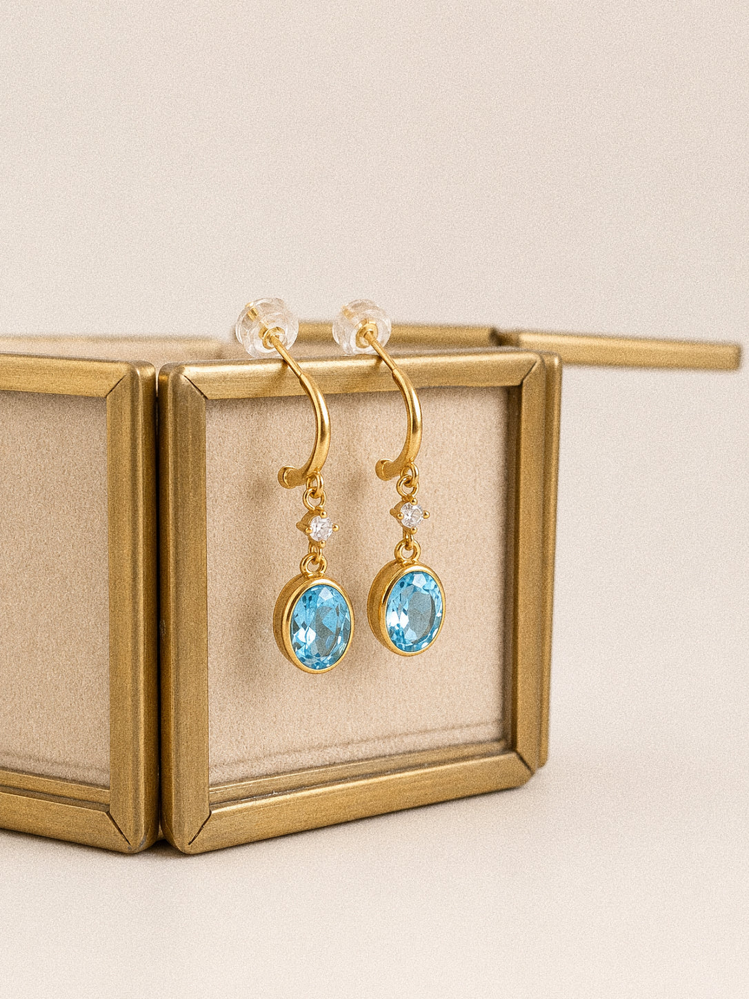 Close view of the Tide gold-plated blue topaz drop earrings showing the oval stones, clear accents and curved posts.