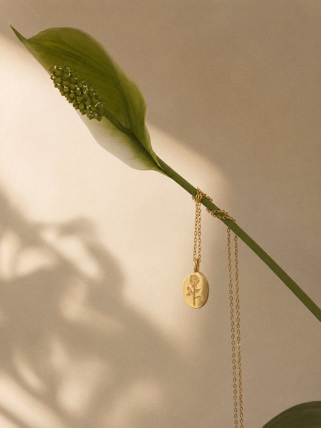 Gold rose pendant necklace styled beside a peace lily flower in warm natural light