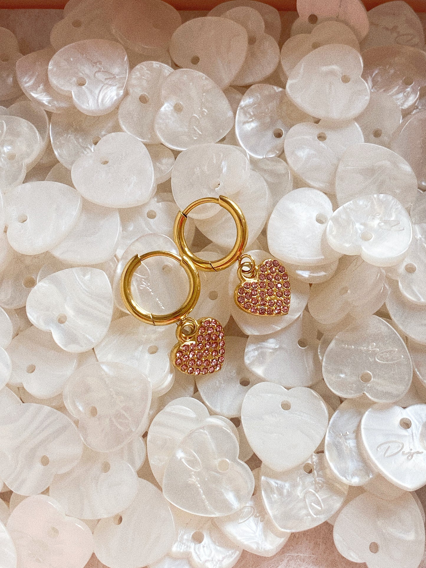 Aesthetic close up of waterproof gold huggie earrings with pink stones resting on white shell hearts.
