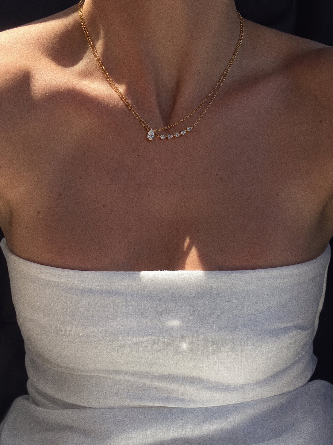 Goutte pear-shaped pendant layered with a curved stone necklace above a white strapless top in sunlight.