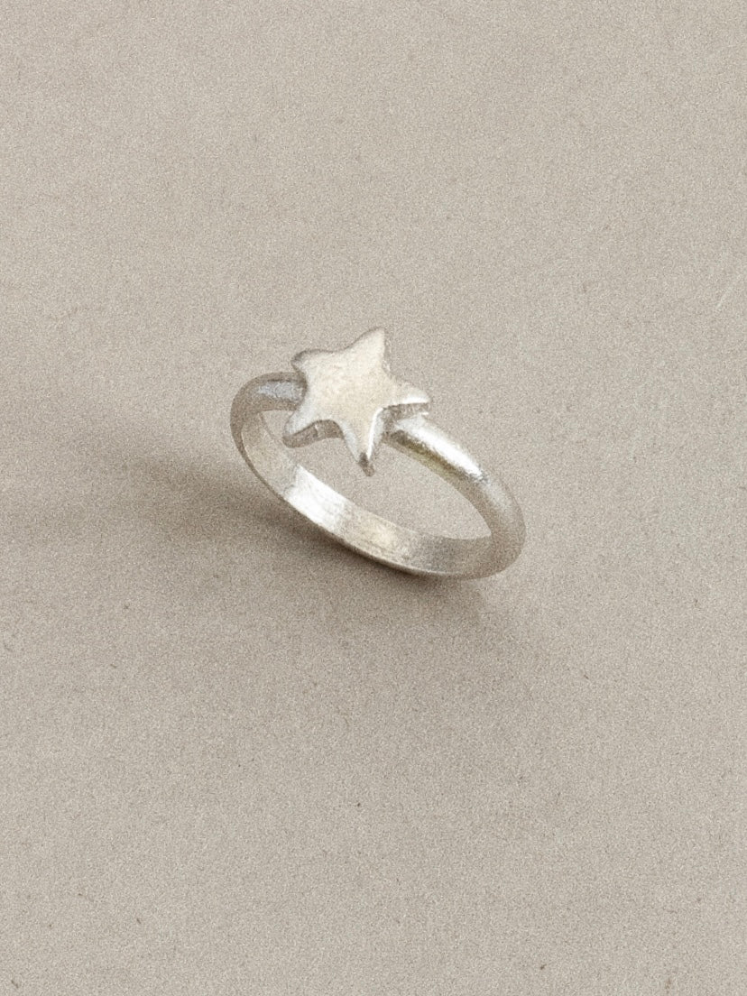 Angled view of a handmade silver star ring on a beige background, brushed star on a rounded 925 band.