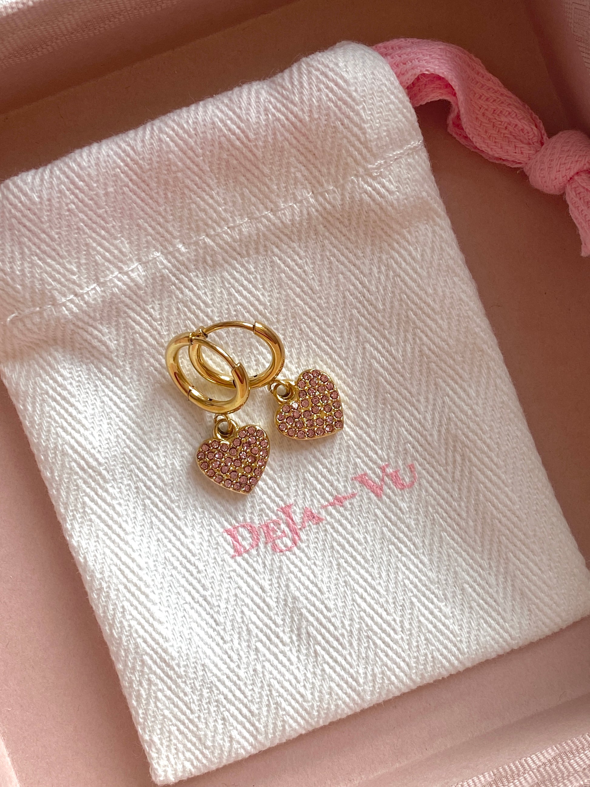 Pink crystal heart earrings displayed on a white Deja vu branded dust bag inside a pink box.