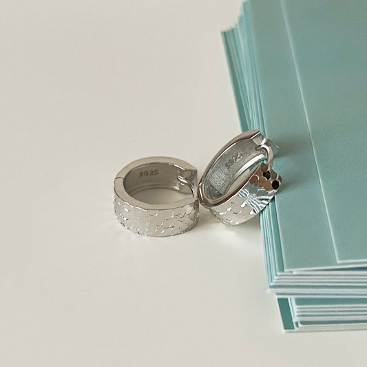 Silver sleeper earrings with diamond-cut texture, resting next to green stationery.