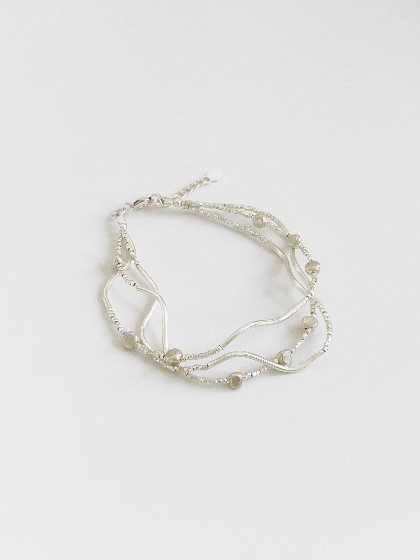 Angled view of a layered silver bracelet mixing three strands of diamond-cut beads, sculptural wave bars and small pebble beads