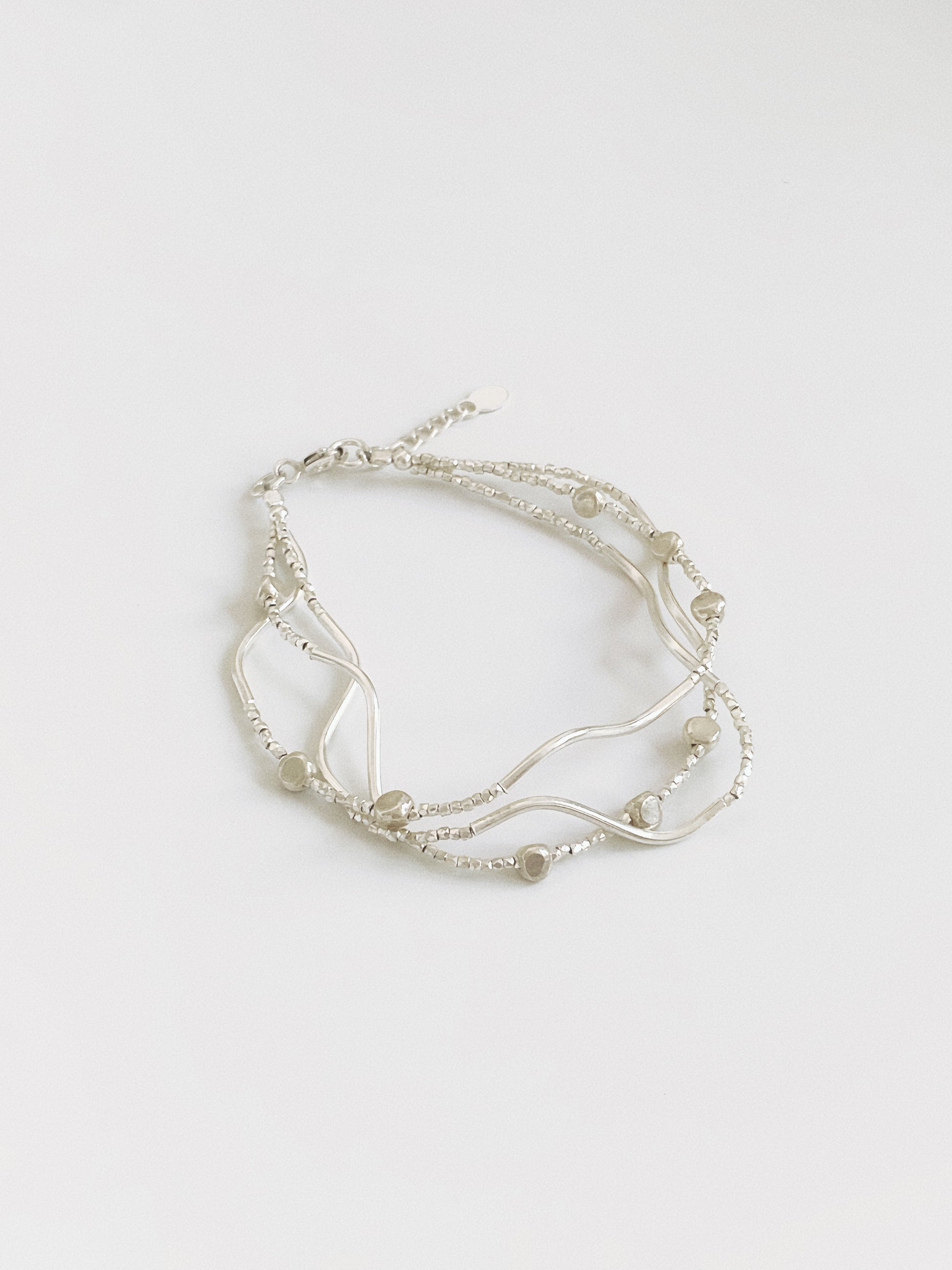 Angled view of a layered silver bracelet mixing three strands of diamond-cut beads, sculptural wave bars and small pebble beads