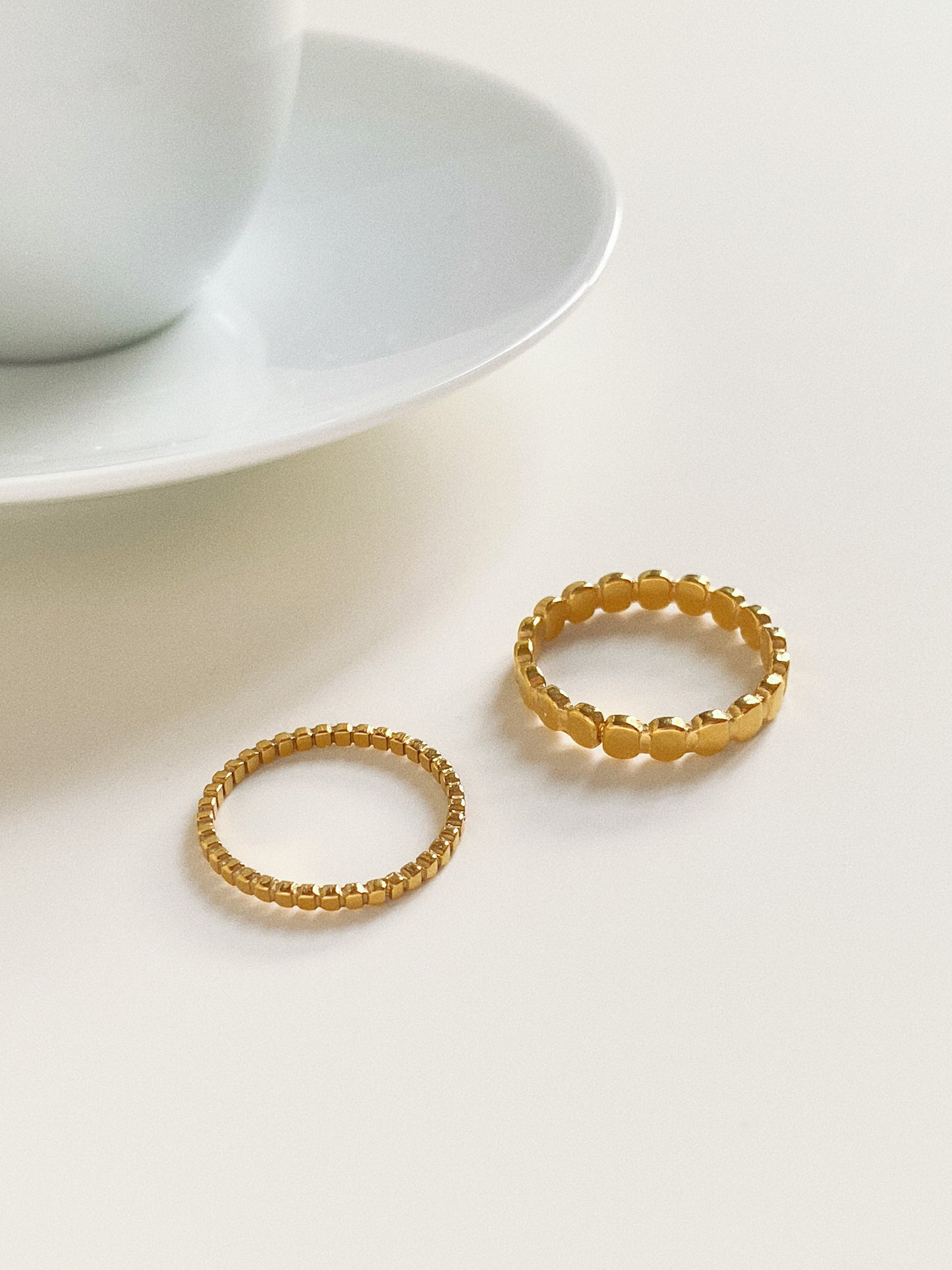 Two minimalist gold beaded rings placed next to a white coffee cup for a sophisticated fashion aesthetic.