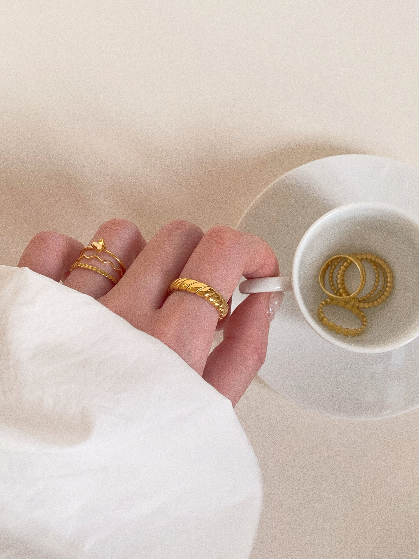 A lifestyle shot of a person holding a white coffee cup, showcasing a stack of gold titanium steel rings including a dainty butterfly design.