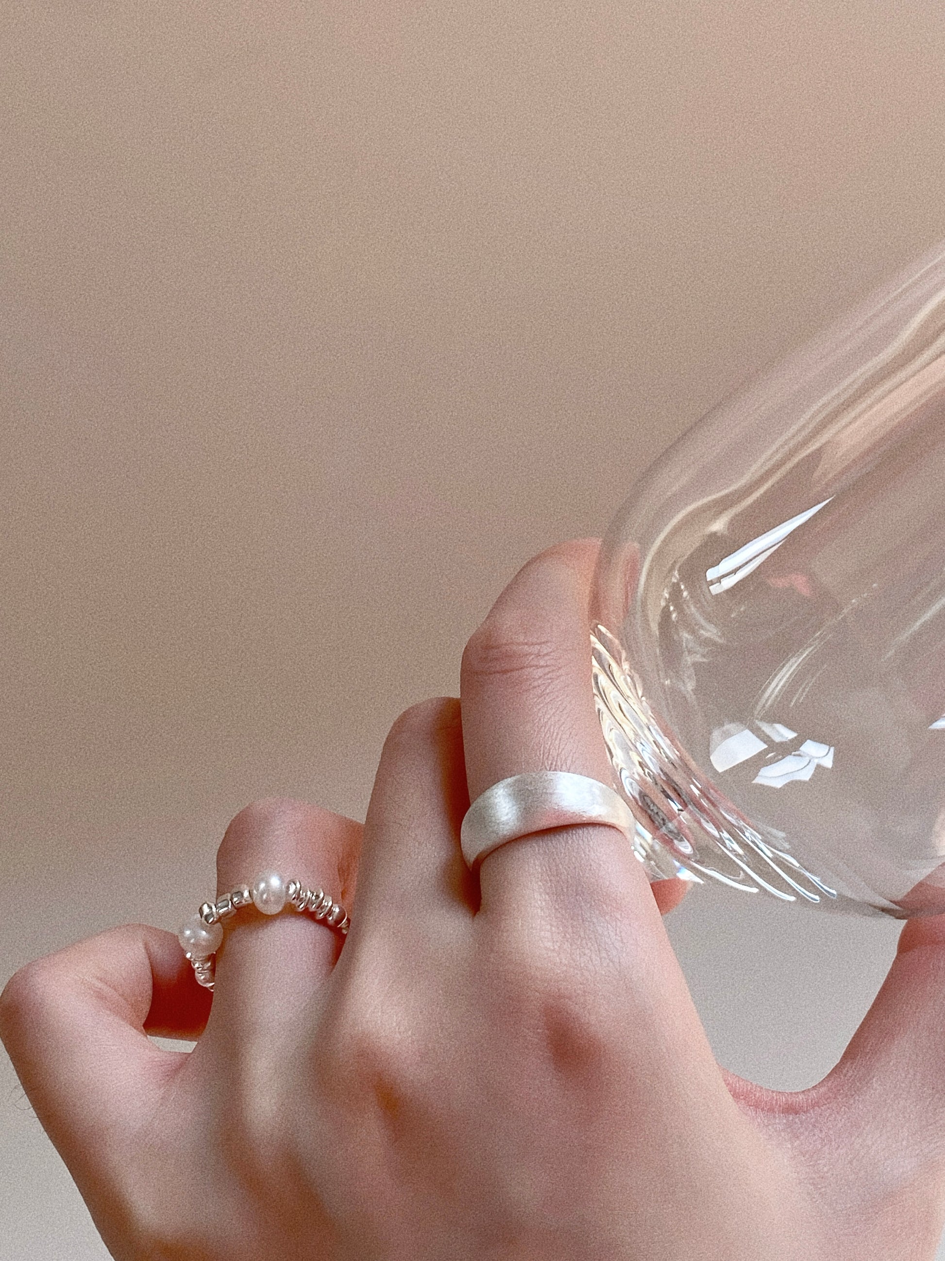 A close-up lifestyle shot of a hand wearing the pearl elastic ring stacked with a thick brushed silver dome ring while holding a clear glass.