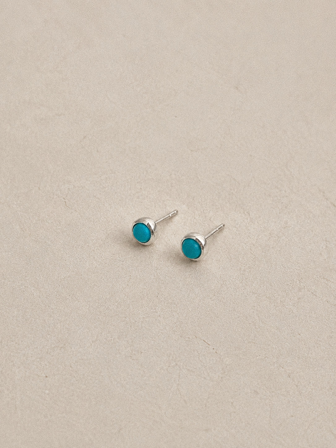 Natural turquoise stud earrings lying on a soft beige backdrop, round blue stones rimmed in polished silver.