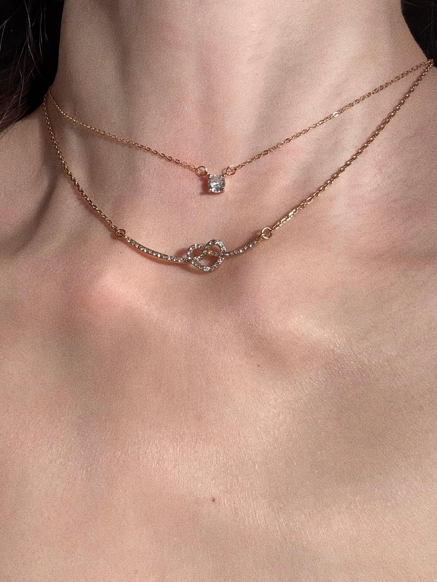 Layered gold necklace stack on neck featuring the knotted heart curved bar necklace paired with a minimalist diamond solitaire choker.