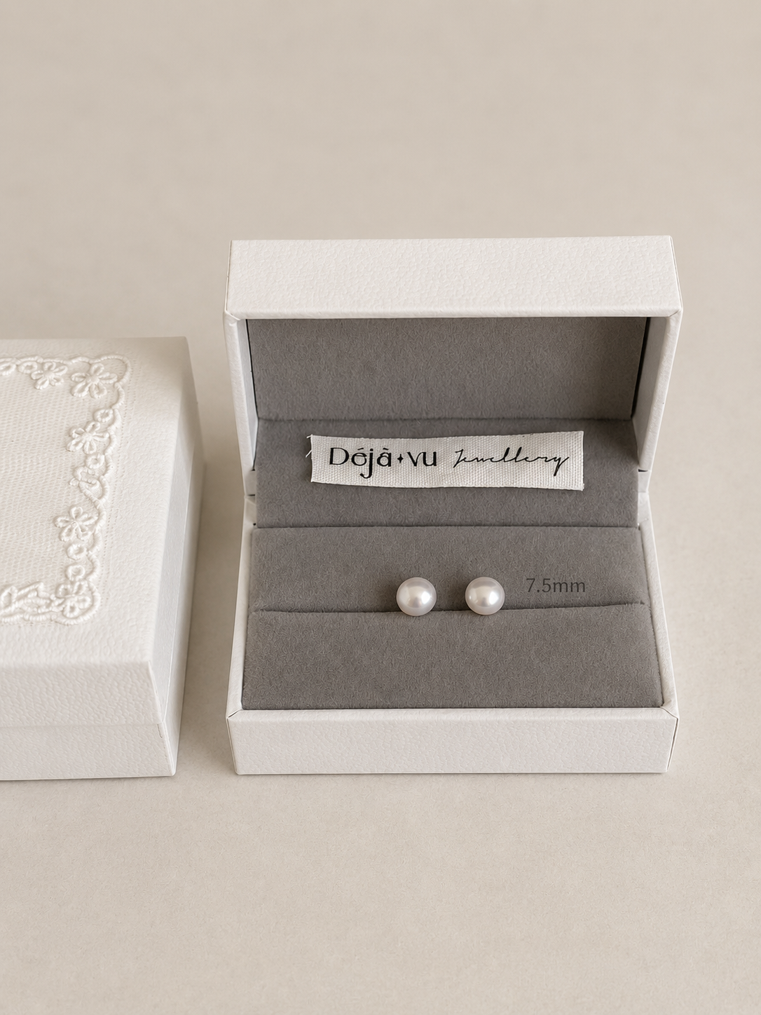 A pair of 7.5 mm white pearl stud earrings in a Deja Vu Jewellery gift box with a stitched fabric brand label.