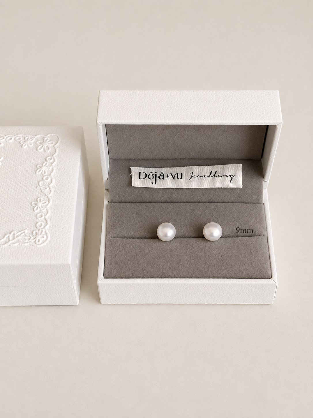 A pair of 9 mm white pearl stud earrings in a Deja Vu Jewellery gift box with a stitched fabric brand label.