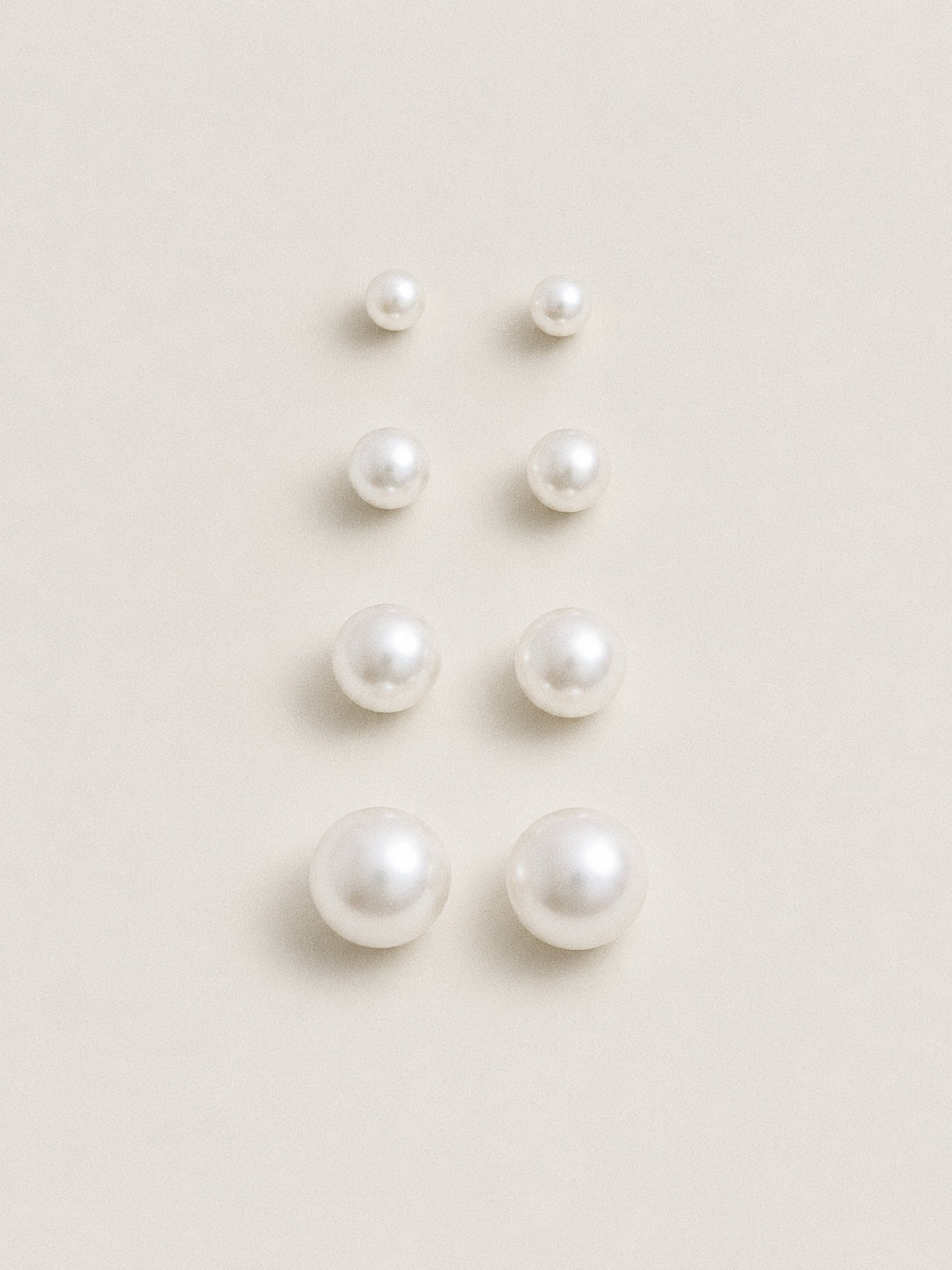 Four pairs of round white pearl stud earrings laid out from smallest to largest on a soft cream backdrop.
