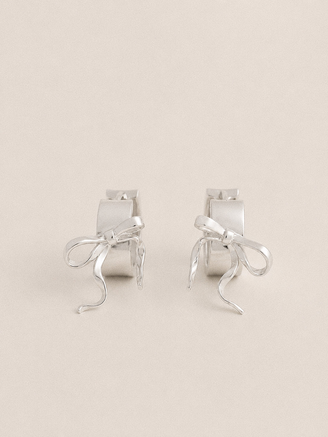 Silver bow huggie hoop earrings with polished trailing ribbon tails, front view on a soft beige backdrop.