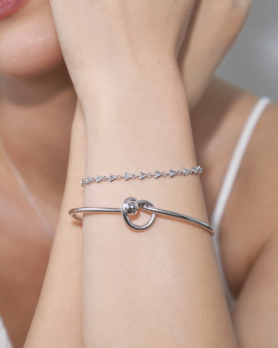 Model wearing a dainty sterling silver heart link bracelet stacked above a silver love knot bangle
