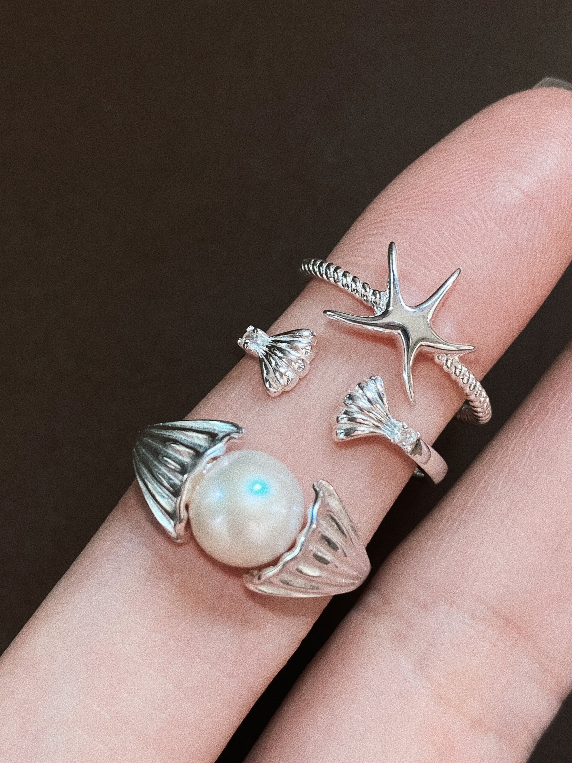 Close-up of a hand wearing a stack of coastal-themed silver rings, including a starfish ring, a shell-shaped ring, and a large pearl shell statement ring.