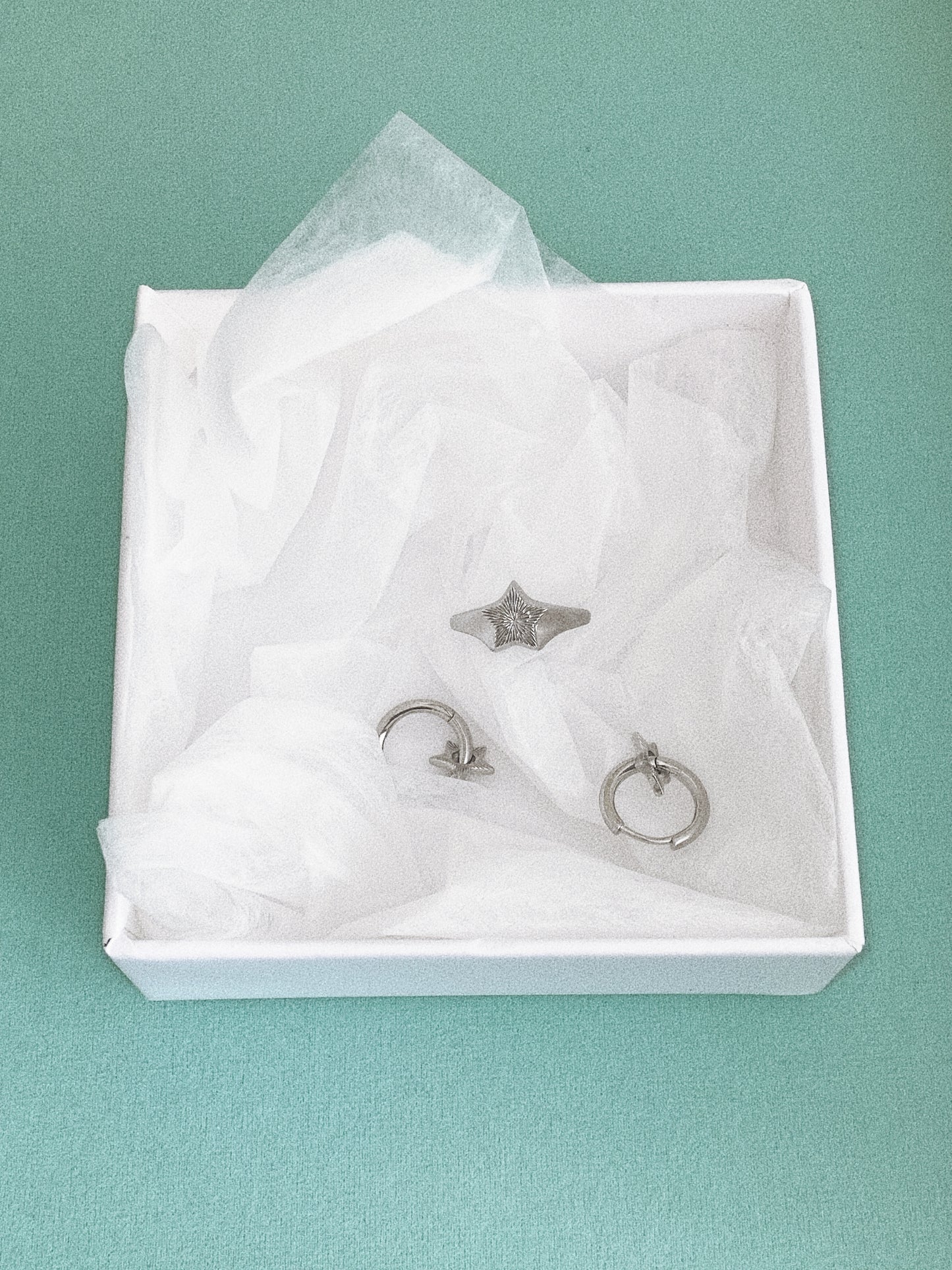 Silver star signet ring in a white box with tissue, shown alongside a pair of star hoop earrings.
