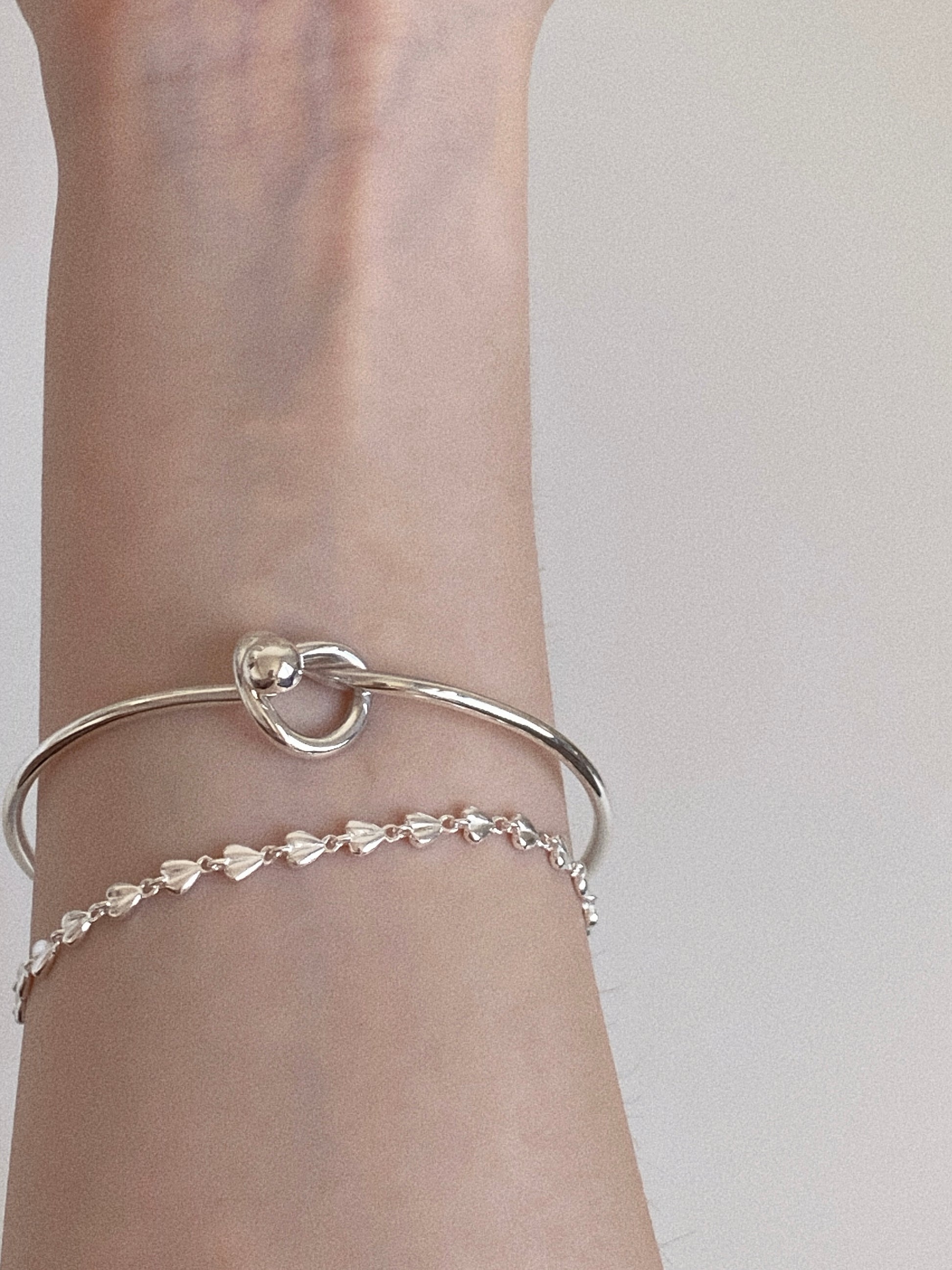 Sterling silver open cuff knot bangle layered with a fine silver heart chain bracelet on a slim wrist