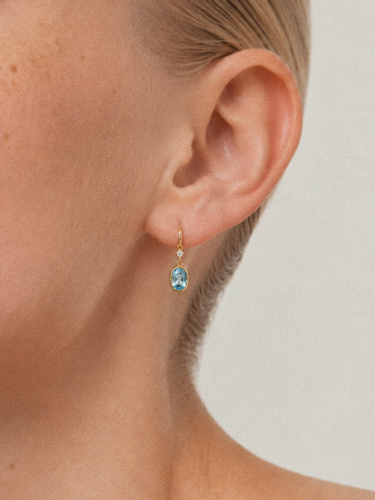 Tide Swiss blue topaz drop earrings worn on the ear, showing their delicate 22 mm length and oval blue stones.