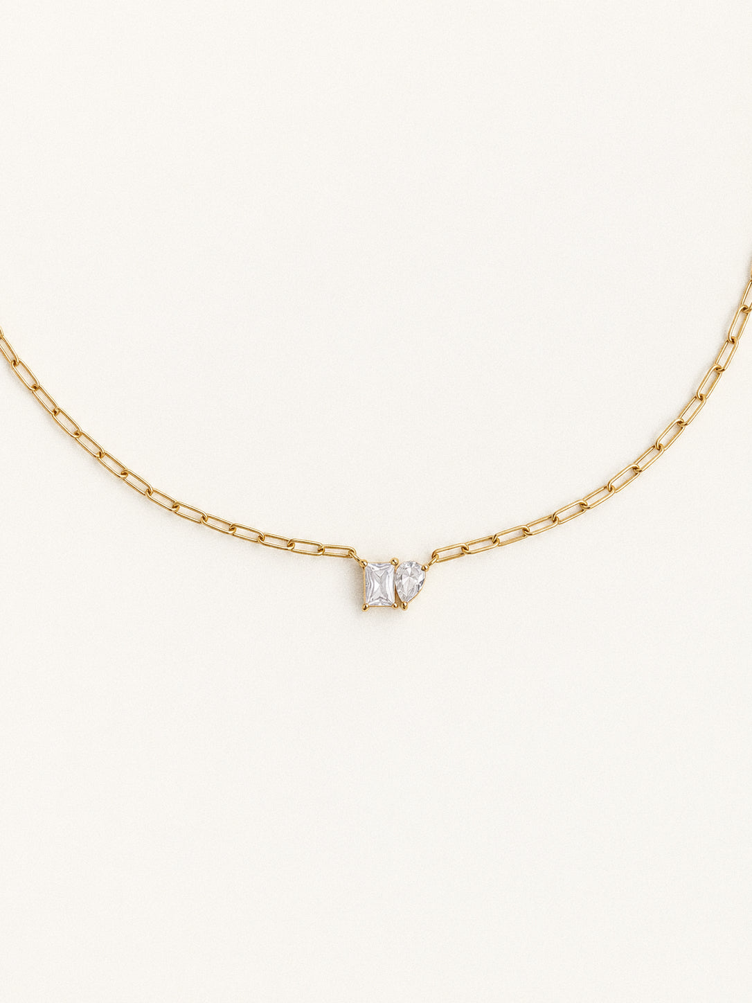 Toi et Moi necklace with rectangular and pear-shaped clear stones on a gold paperclip chain against an ivory background.