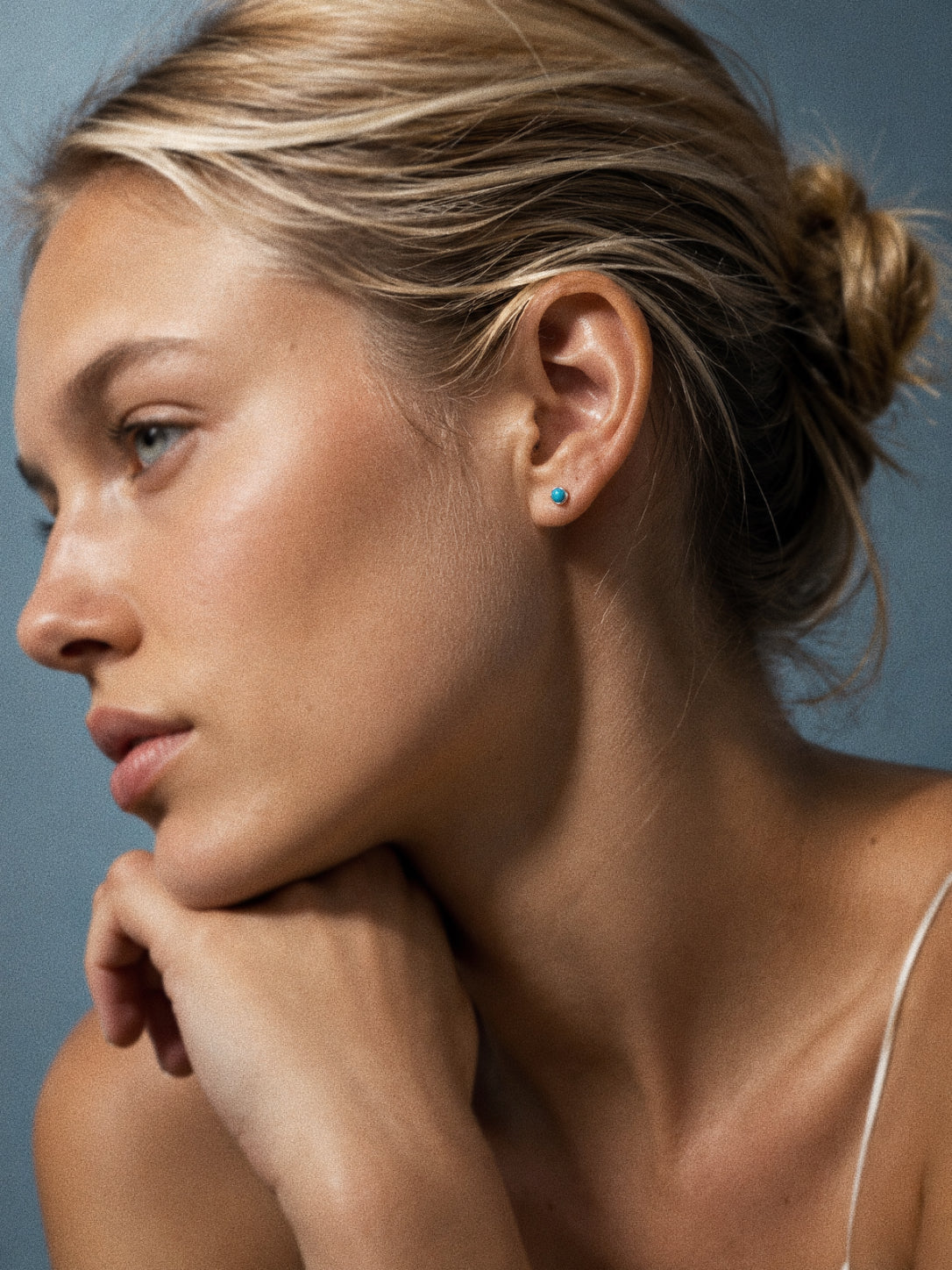 Turquoise stud earring worn in the ear lobe with a white slip top against a soft blue backdrop.